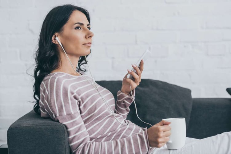 Attractive pensive woman listening music in earphones and looking away while resting on sofa
