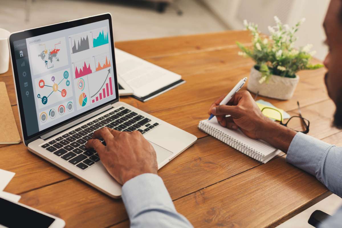 Close up back view of black entrepreneur analyzing graph on laptop and writing in notepad