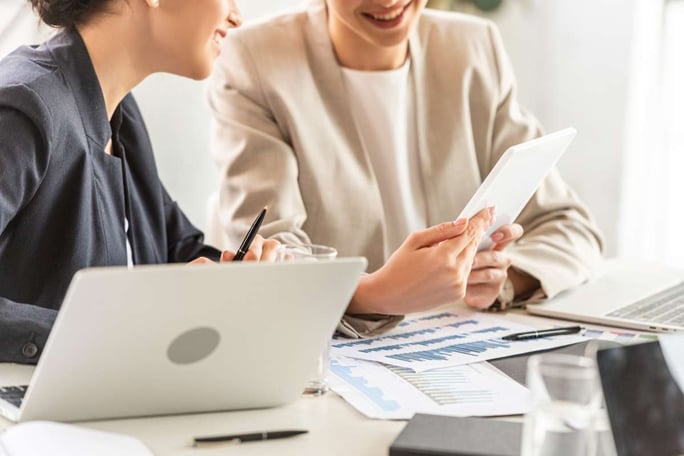 Partial view of two businesswomen in formal wear at table with laptops and digital tablet in office