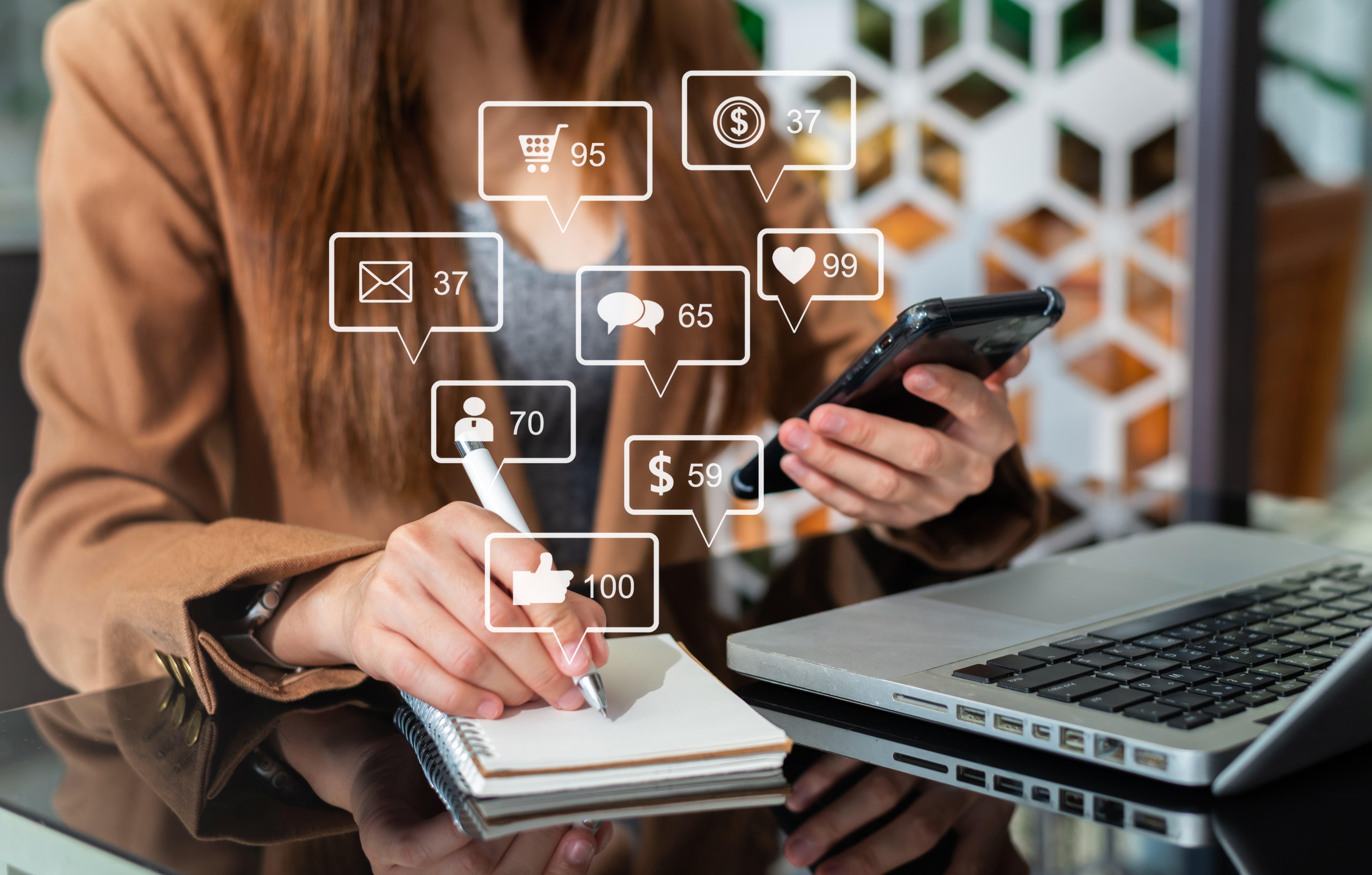 Image of lady holding her phone while in front of her laptop and writing down notes