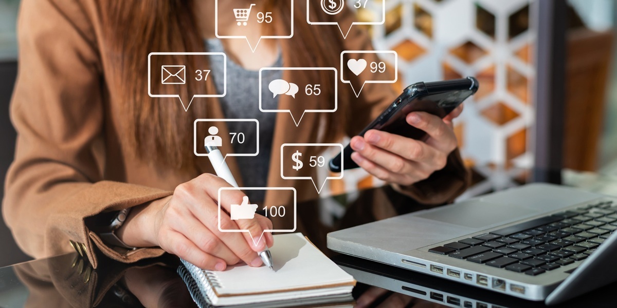 Image of lady holding her phone while in front of her laptop and writing down notes