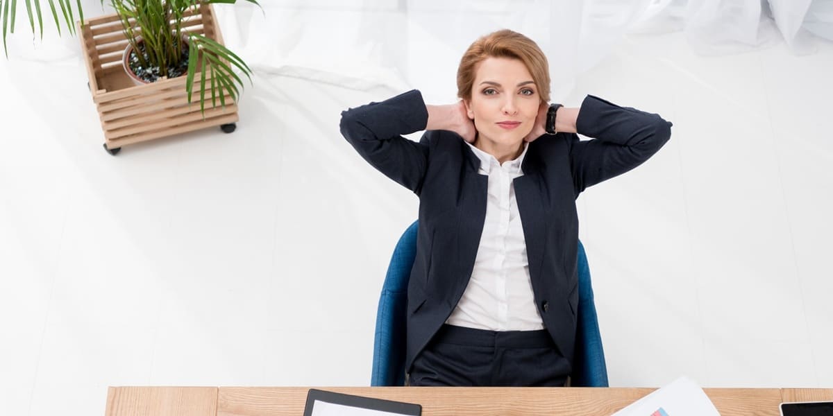 Woman in a suit seated on her desk smiling and with her hands on her head
