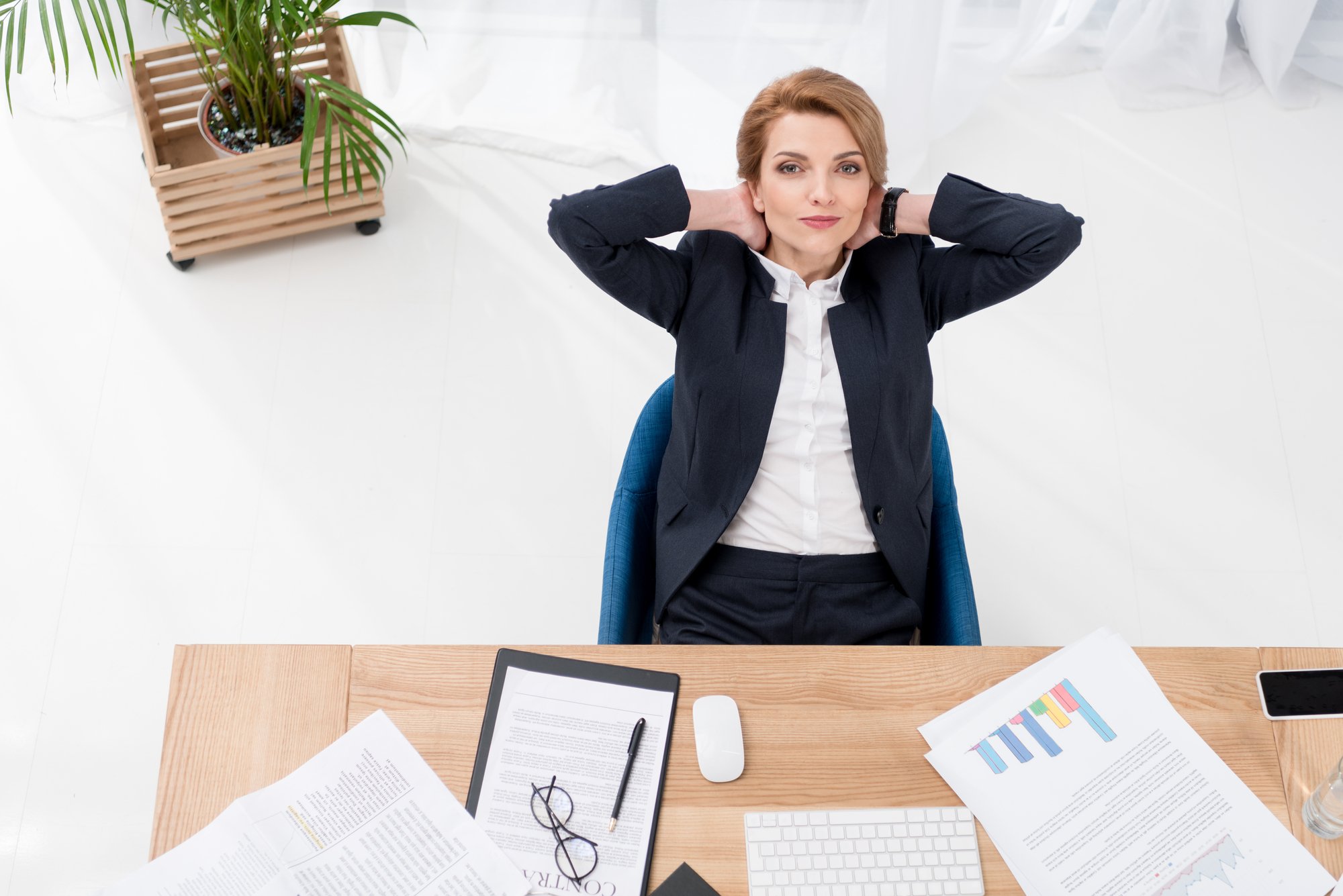 Woman in a suit seated on her desk smiling and with her hands on her head